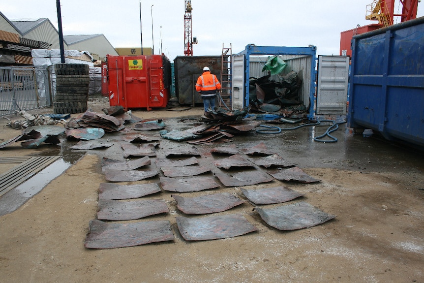 At an industrial site, you see rows of copper plates dating back to the medieval-era, with coloured shipping containers behind.
