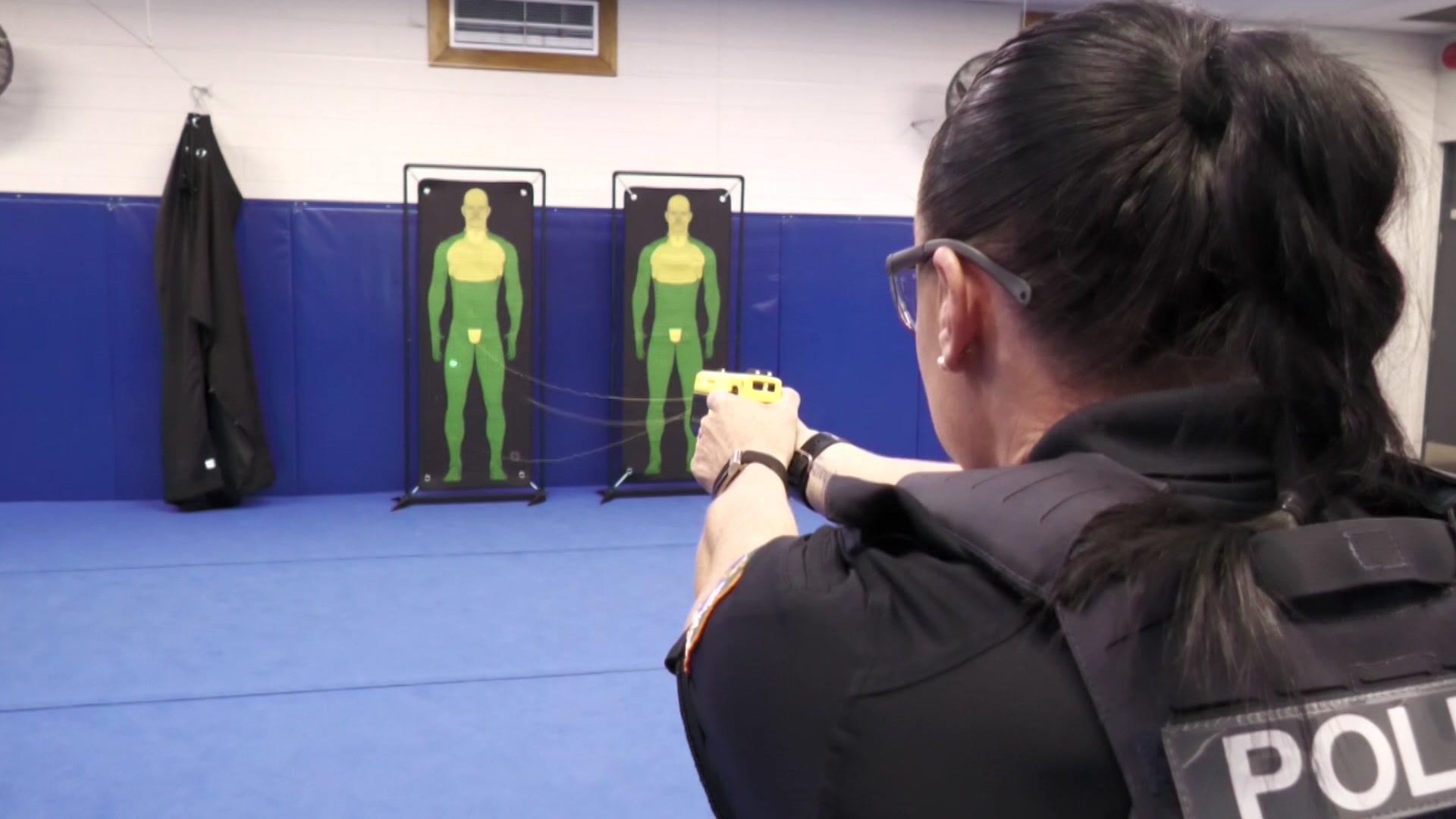 A uniformed police officer fires a taser device, striking a dummy standing against the wall of a training facility.