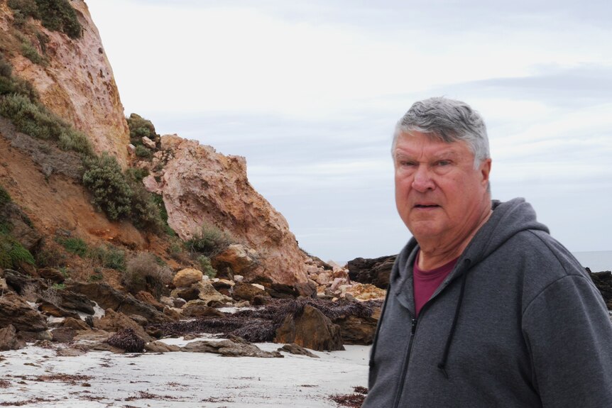 Man in foreground looking at camera, standing near collapsed cliff, ocean and rocks in background