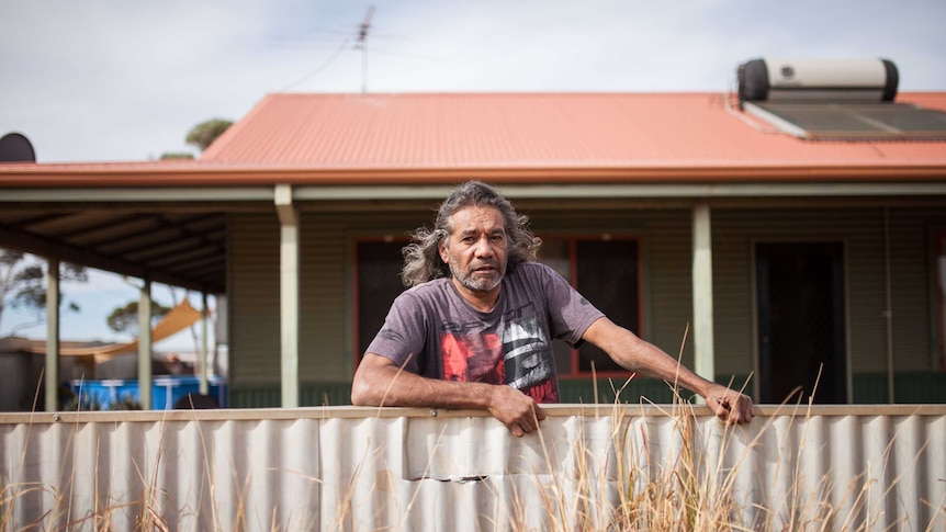 A man stands at a broken fence outside his home