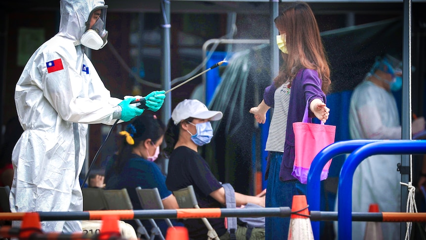 A woman in a face mask stands with arms outstretched as a man in full PPE sprays her with disinfectant
