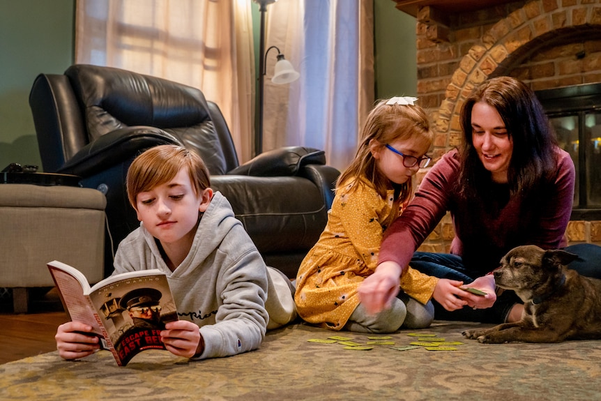 A boy in a grey hoodie reads a book in front of the fireplace next to a girl in a yellow dress, a woman and a dog