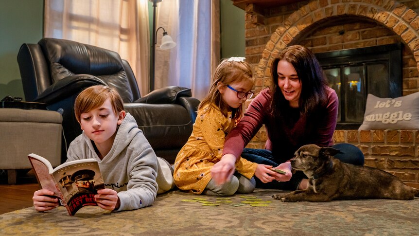 A boy in a grey hoodie reads a book in front of the fireplace next to a girl in a yellow dress, a woman and a dog