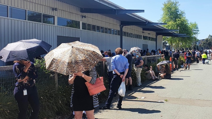 Dozens of people are seen lining up for coronavirus testing next to a building in Shepparton, some are standing under umbrellas.