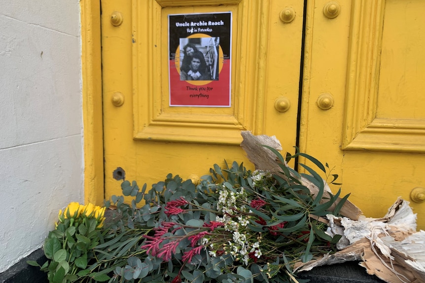 Flowers wrapped in paperbark outside a yellow door, with a tribute to Archie Roach poster behind.