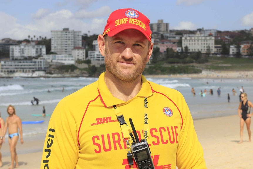 A lifeguard, wearing his full yellow and red kit, poses for a photo with Bondi Beach in the background.
