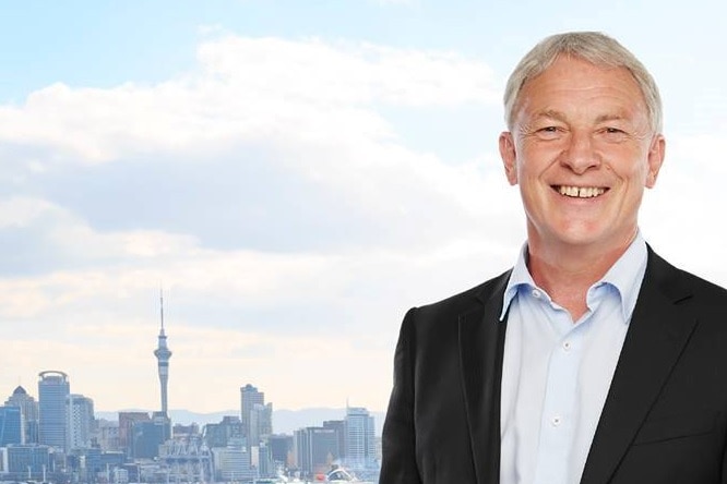 Man in a suit smiles in front of the Auckland skyline