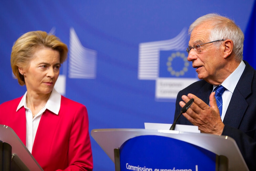 Two people stand behind lecterns in the blue of the EU flag in front of lecterns with the EU flags.