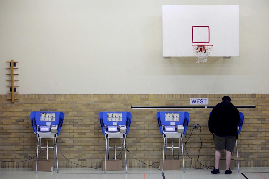 A man stands at one of four electronic voting machines. Above him is a basketball hoop.