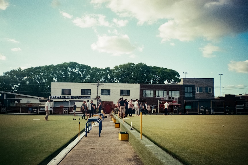Men assemble on two bowling greens with a two-storey clubhouse in the background