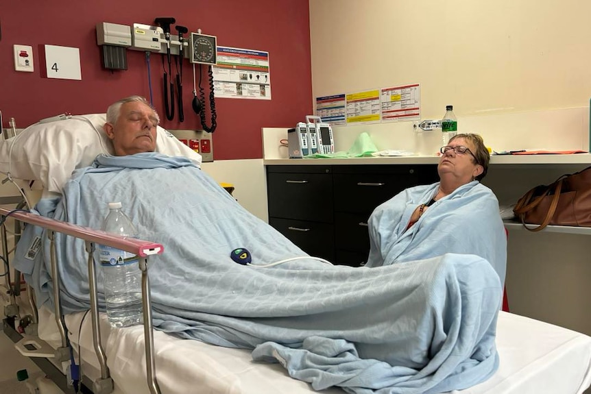 a man sleeps in an emergency department hospital bed wrapped in a blue blanket. a woman sleeps sitting up on a chair beside