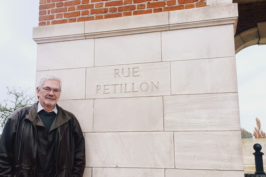 A man has short grey hair, he wears a brown leather coat, behind him is white sandstone with Rue Petillon written