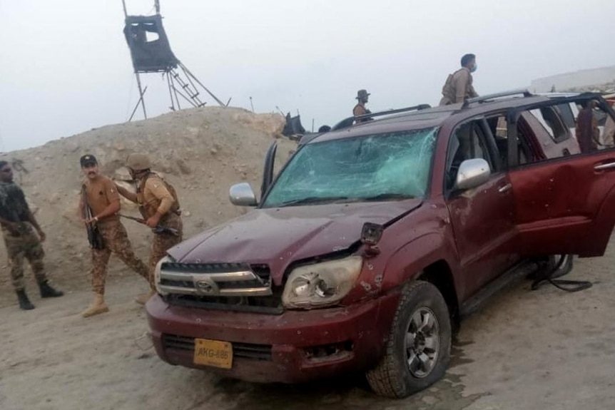 A damaged maroon-coloured four-wheel drive sits on a dusty road with armed men in khaki standing behind.