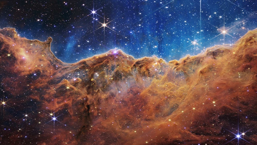 Orange, mountainous clouds in a starry sky. This image shows an area of star birth, taken by NASA’s James Webb Space Telescope.
