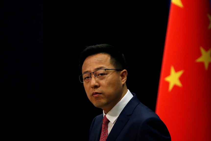 A man in a suit standing in front of the Chinese flag looks pensively towards the camera
