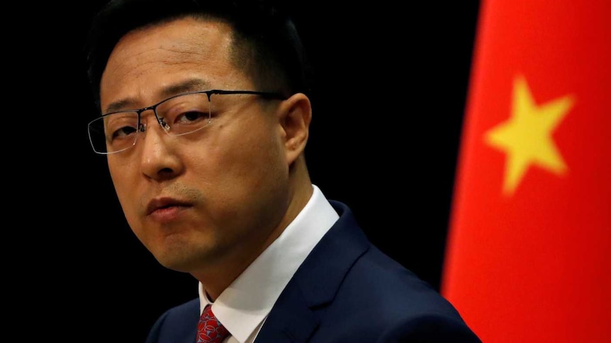 A man in a suit standing in front of the Chinese flag looks pensively towards the camera