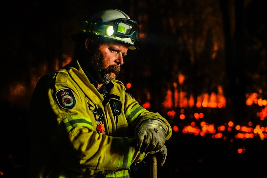 A firefighter stands with his hands on an axe as he stares into the distance. It is dark and the torch on his helmet is on.