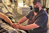 A man wearing a mask holds the hand of a man in hospital bed. 