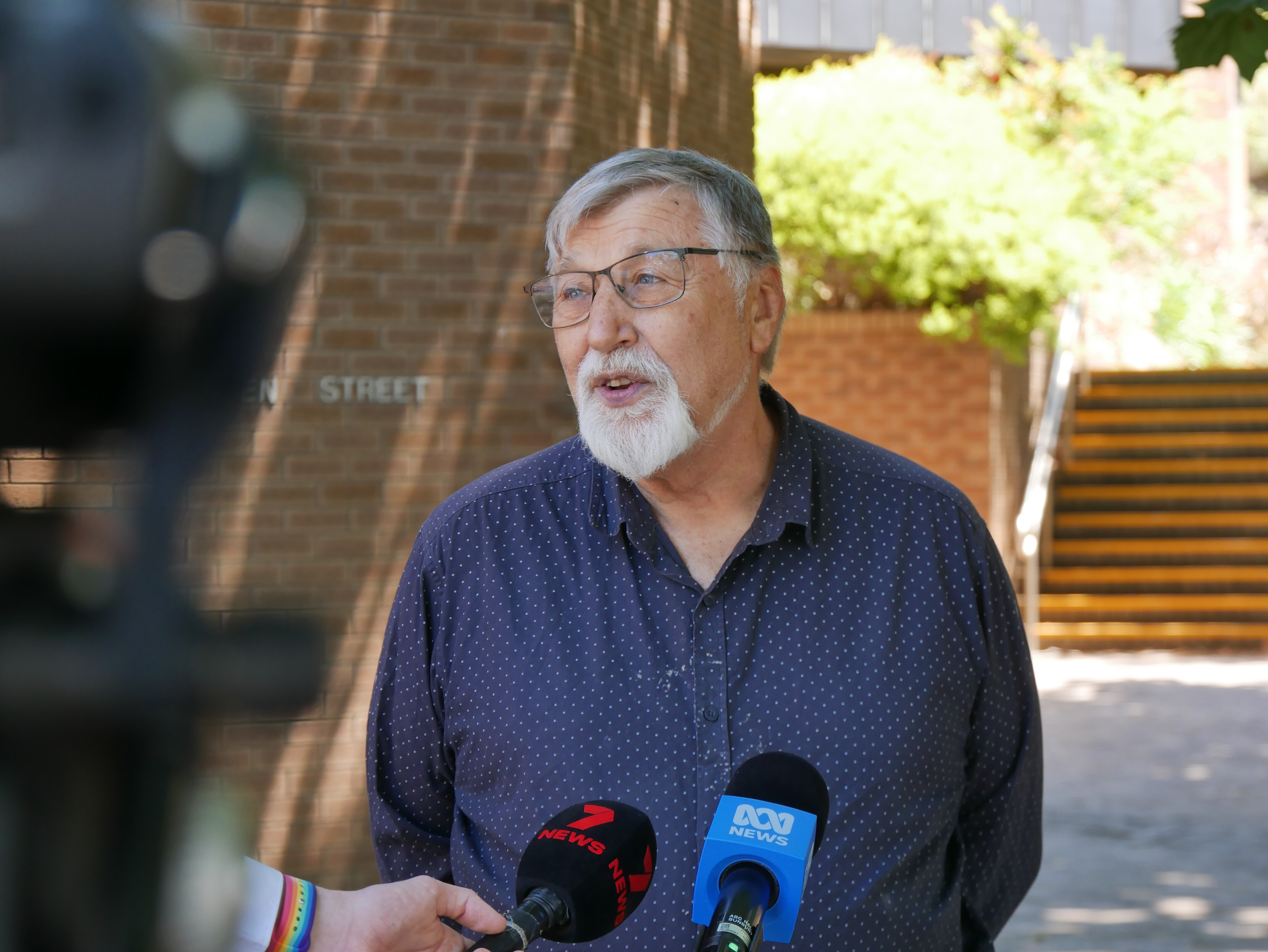 Un hombre con pelo y barba grises, vestido con una camisa azul oscuro, habla con los medios 