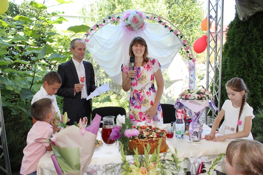 A man and woman stand surrounded by children at a table decorated with flowers. 