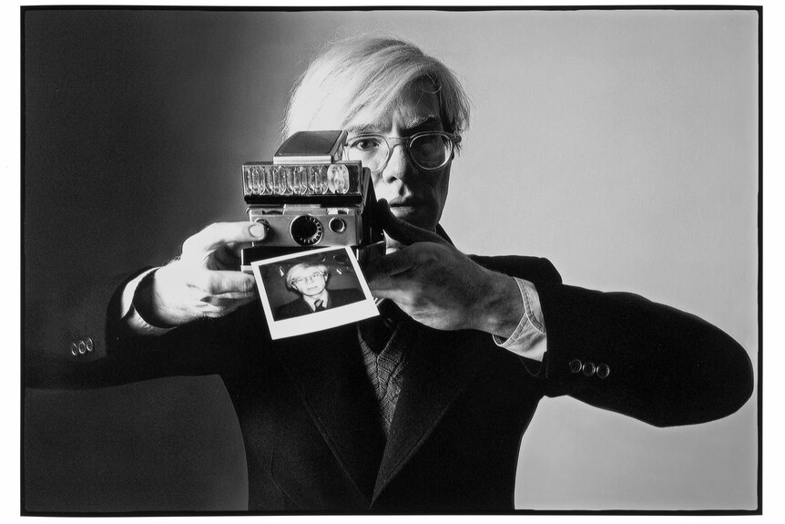 A black-and-white photograph of Andy Warhol moving a polaroid camera away from his face. The polaroid is his portrait.