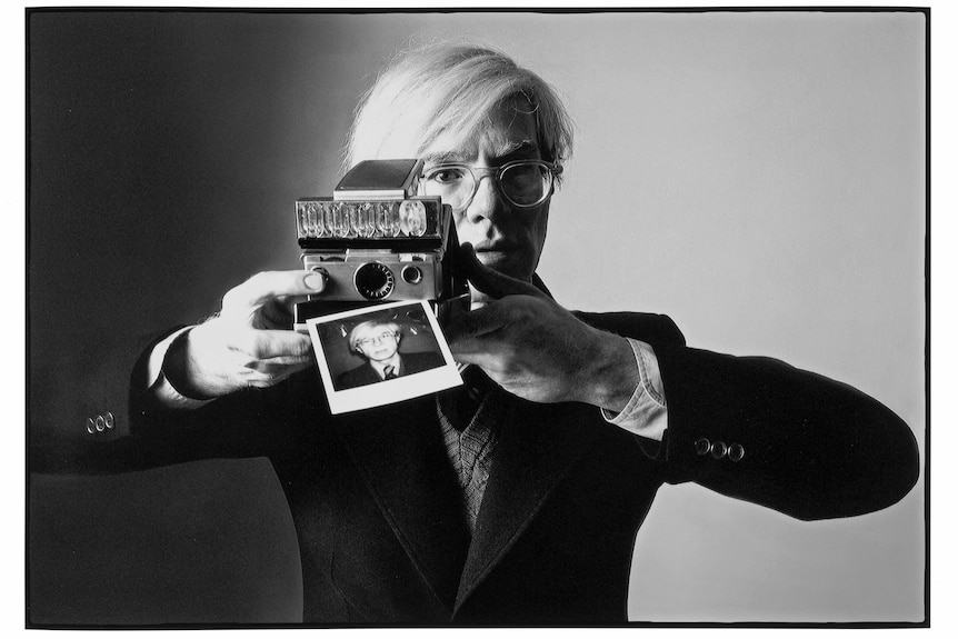 A black-and-white photograph of Andy Warhol moving a polaroid camera away from his face. The polaroid is his portrait.