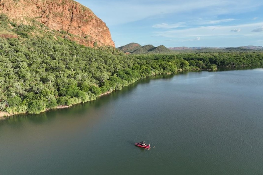 a drone shot of a ski boat in a wide river next to green trees and an orange ridge