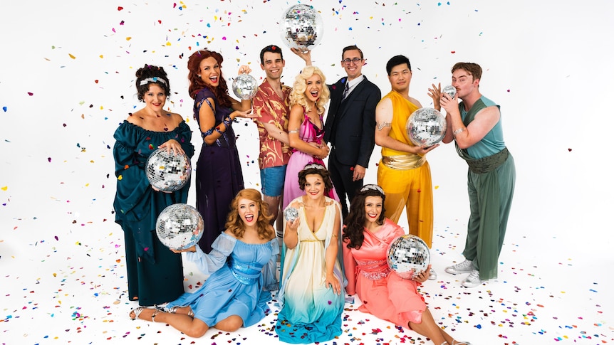 A group of people stand and sit holding disco balls in colourful costumes with glitter in the air in a white studio space.