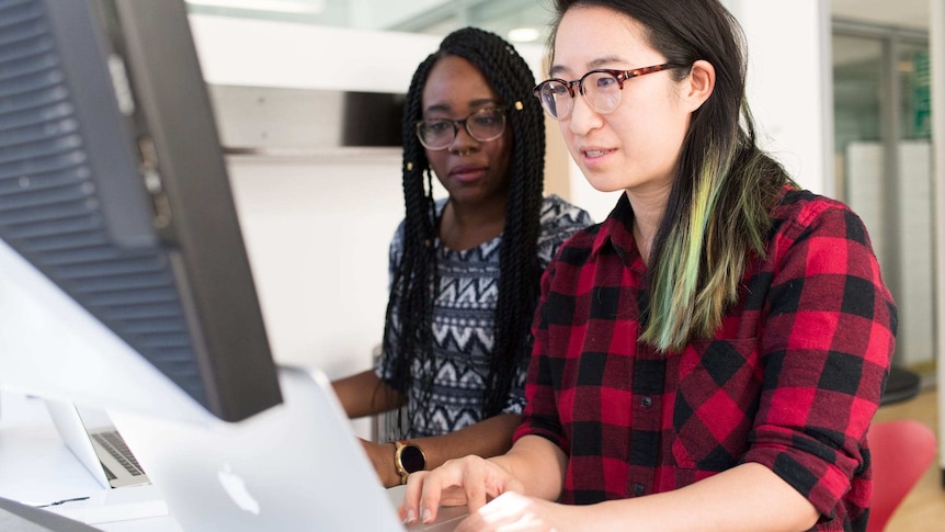 Two woman are focused on a computer screen at a desk.