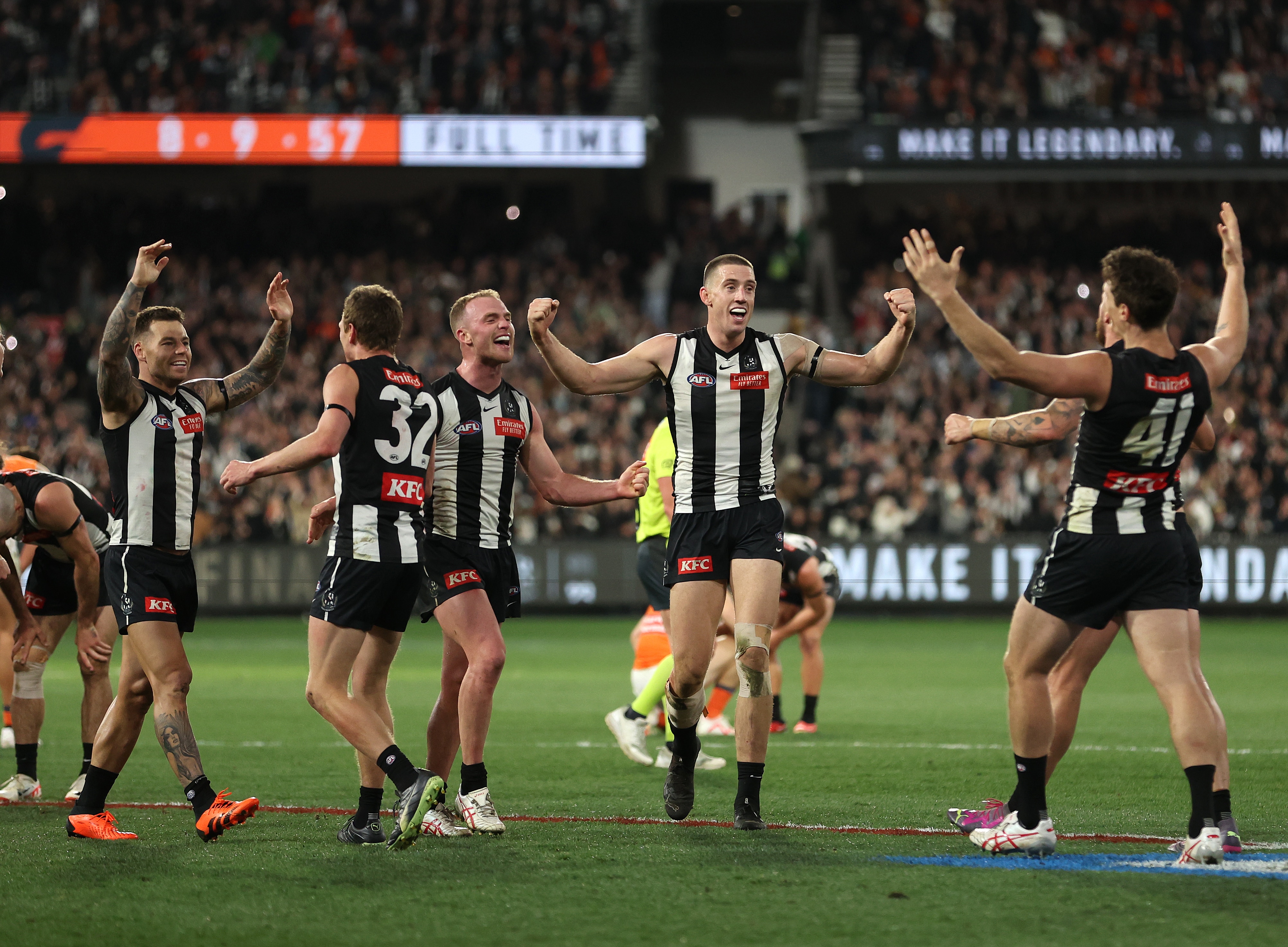 A group of Collingwood AFL players stand with arms raised in triumph after the final siren in a finals game.
