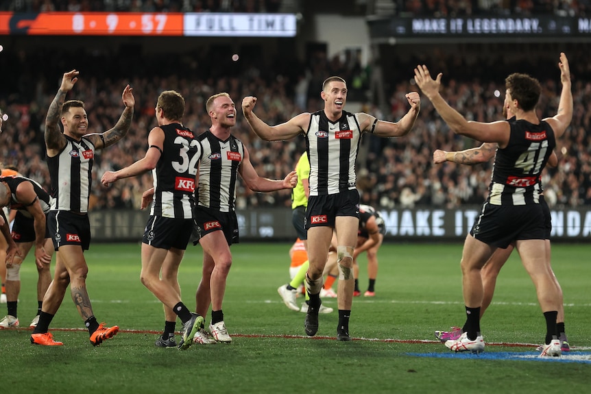 A group of Collingwood AFL players stand with arms raised in triumph after the final siren in a finals game.