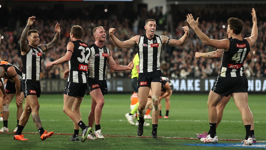 A group of Collingwood AFL players stand with arms raised in triumph after the final siren in a finals game.