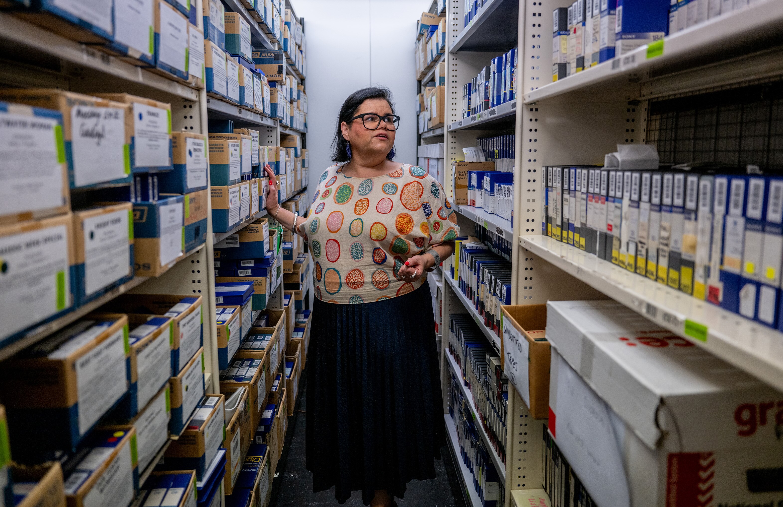 Dark haired woman wutg glasses in long skirt and white shirt with colourful circles stands between shelves with archive material