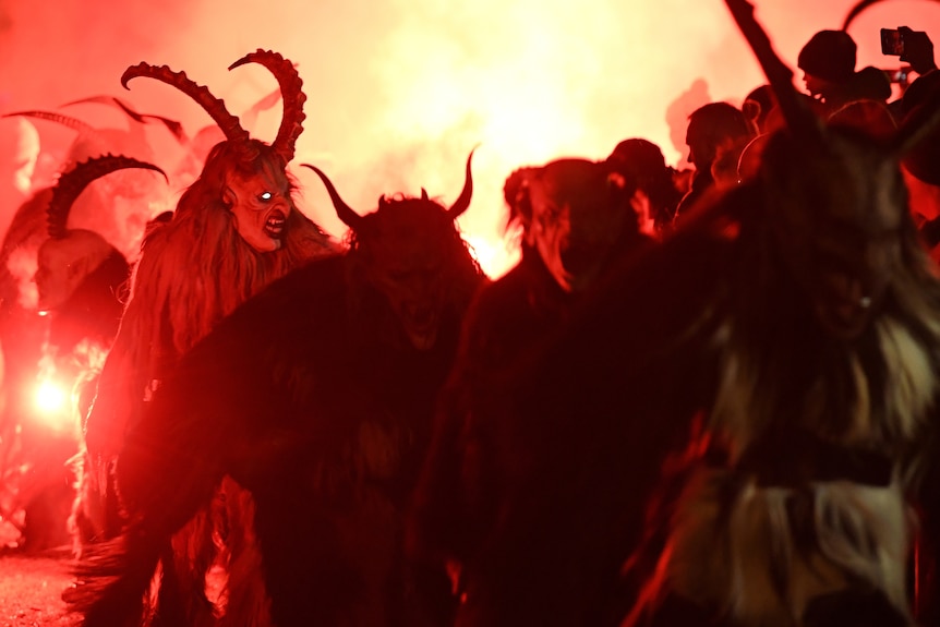 People in traditional goat demon masks run past a crowd of onlookers while holding red flares.