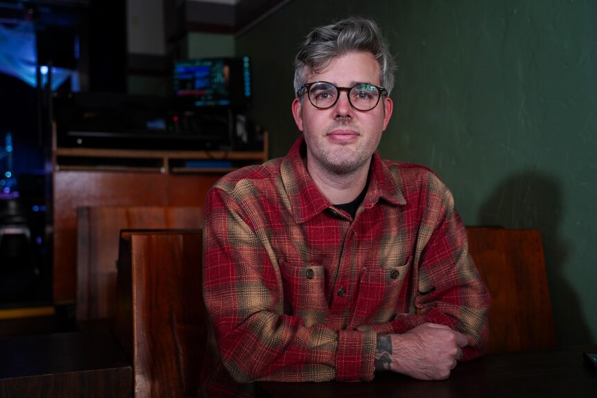 A man with greying hair, dark-rimmed glasses and a red flannel shirt poses for a photo sitting down indoors.