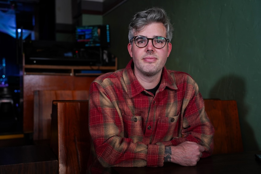 A man with greying hair, dark-rimmed glasses and a red flannel shirt poses for a photo sitting down indoors.