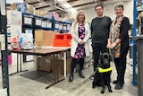Elisa, Adam and Annette standing in warehouse. Annette is holding the lead of a black Labrador guide dog 
