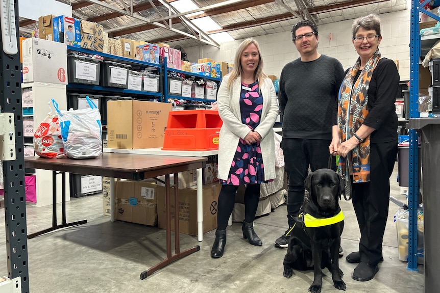 Elisa, Adam and Annette standing in warehouse. Annette is holding the lead of a black Labrador guide dog 