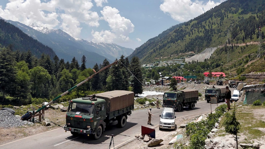 A convoy of three army trucks stops at a control point on a paved road running through a verdant valley.
