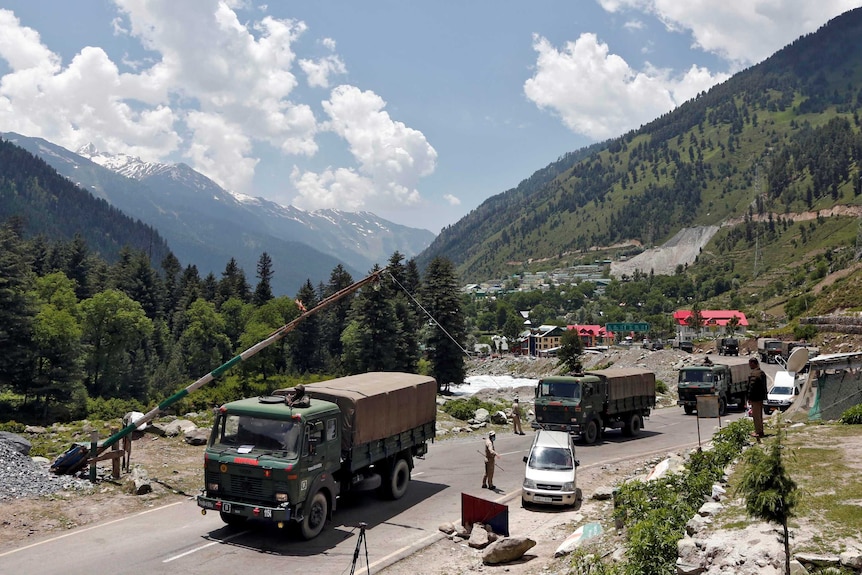 A convoy of three army trucks stops at a control point on a paved road running through a verdant valley.