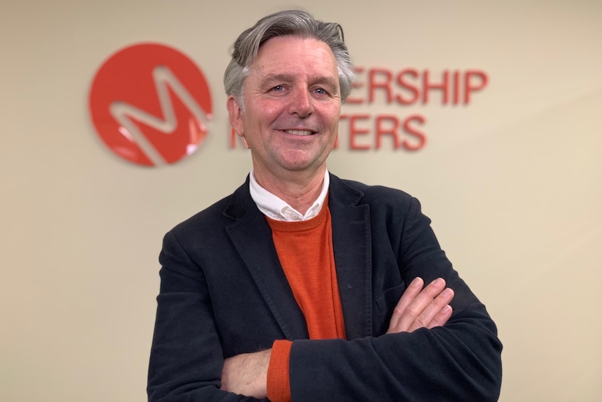 A middle-aged, grey-haired man in black jacket, red jumper & white shirt stands in front of a company logo on an interior wall