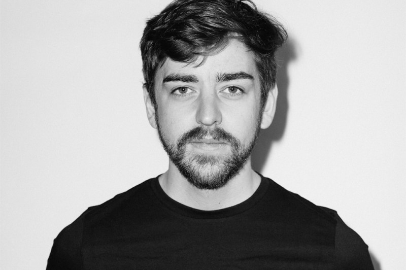 A black-and-white photo of a young man with a beard posing formally for a photo, and not smiling.