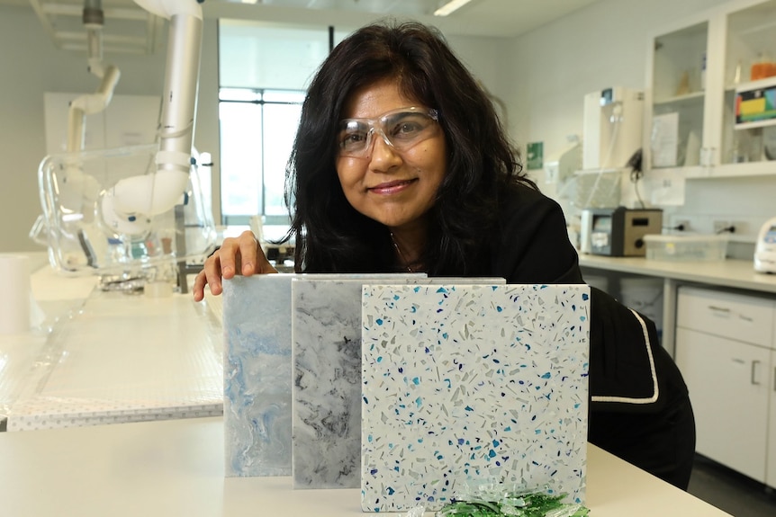A woman wearing clear safety glasses smiles in a lab next to three marble-like tiles.
