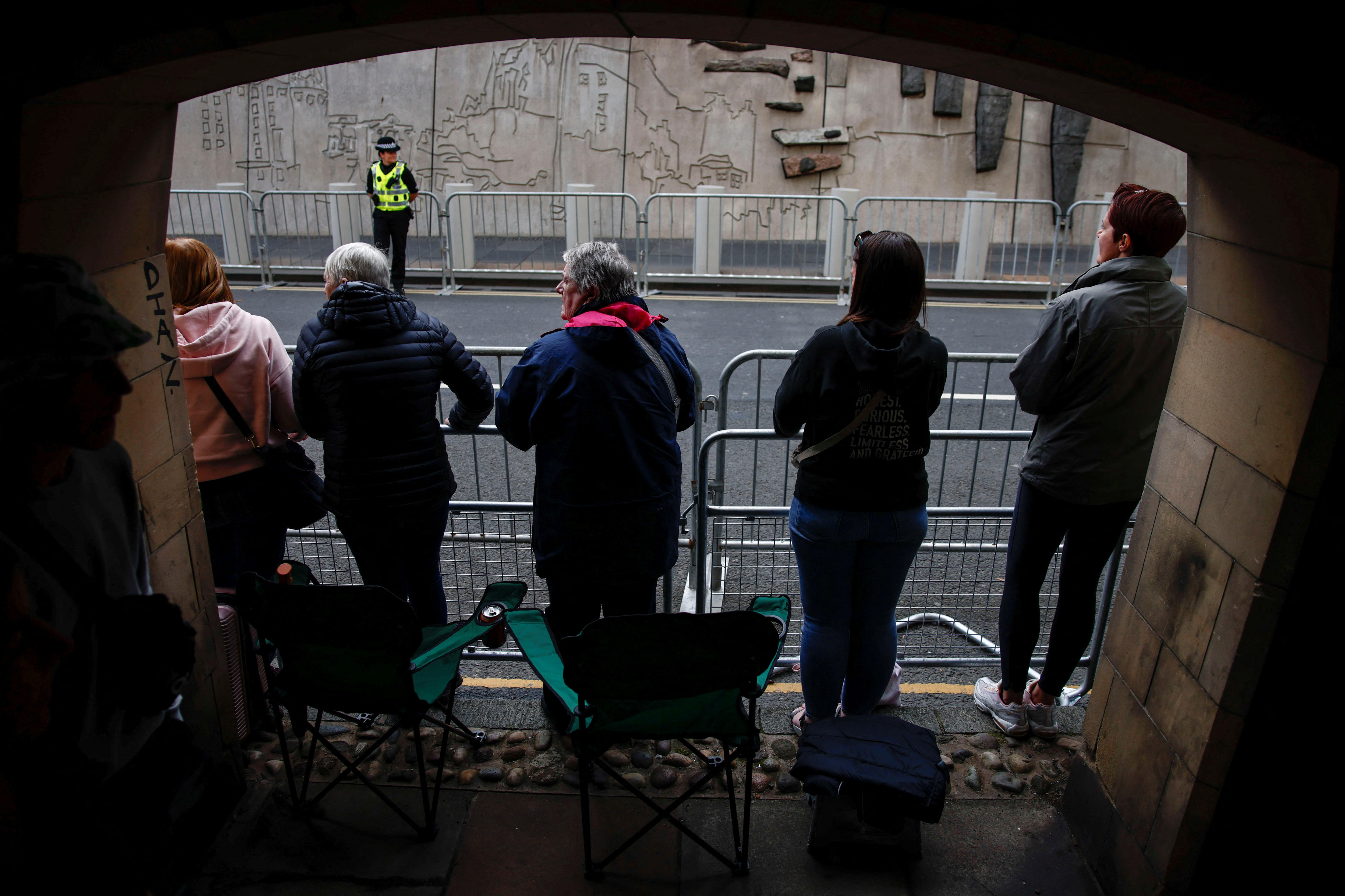People lean on a barricade beside a road, with camping chairs behind them. 