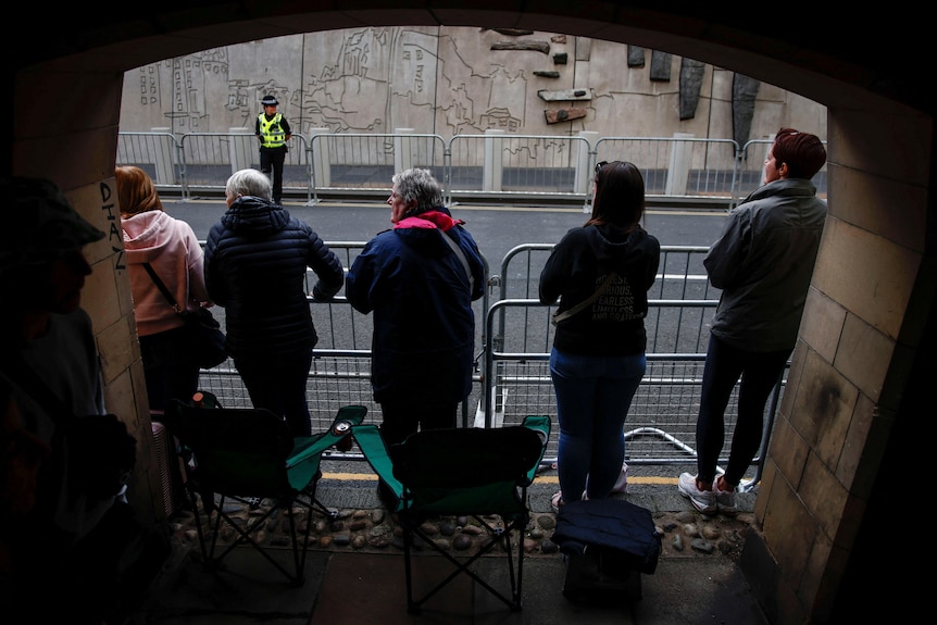 People lean on a barricade beside a road, with camping chairs behind them. 