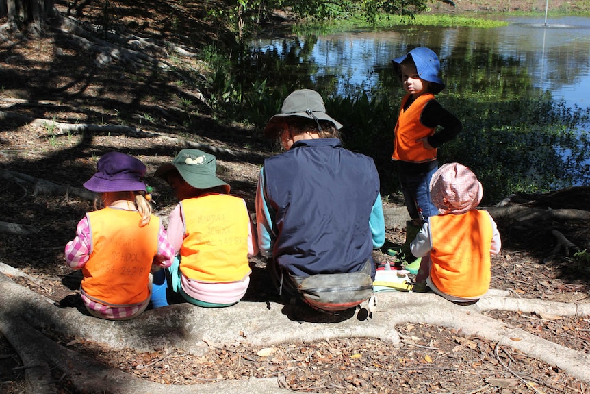 A group of Nature School students gather for lunch on a log by a waterway.