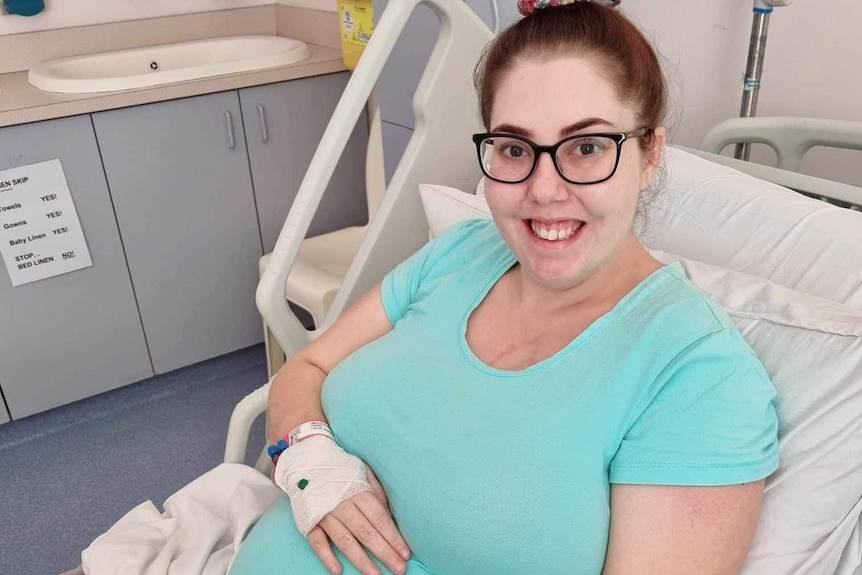 A woman lying in a hospital bed pregnant and smiling at the camera. 