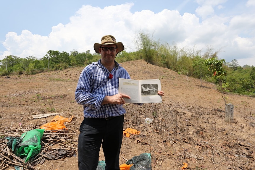 A man in a broad-brimmed hat stands holding a historic photo on a bare hill rising behind him
