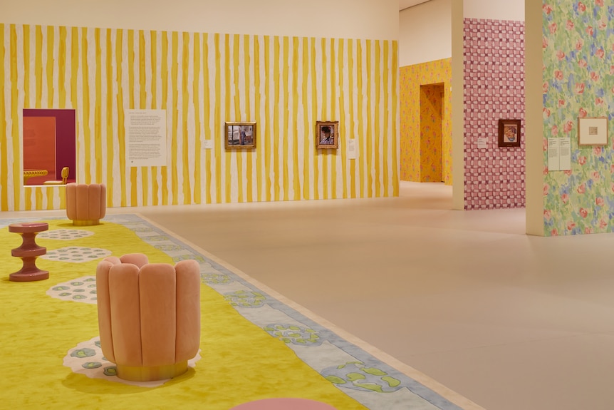 A spacious room inside a gallery featuring a yellow striped feature wall, and velvet-like peach chairs.