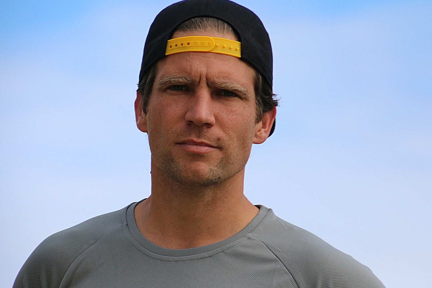 A headshot of a man in a grey t-shirt and backwards black baseball cap.